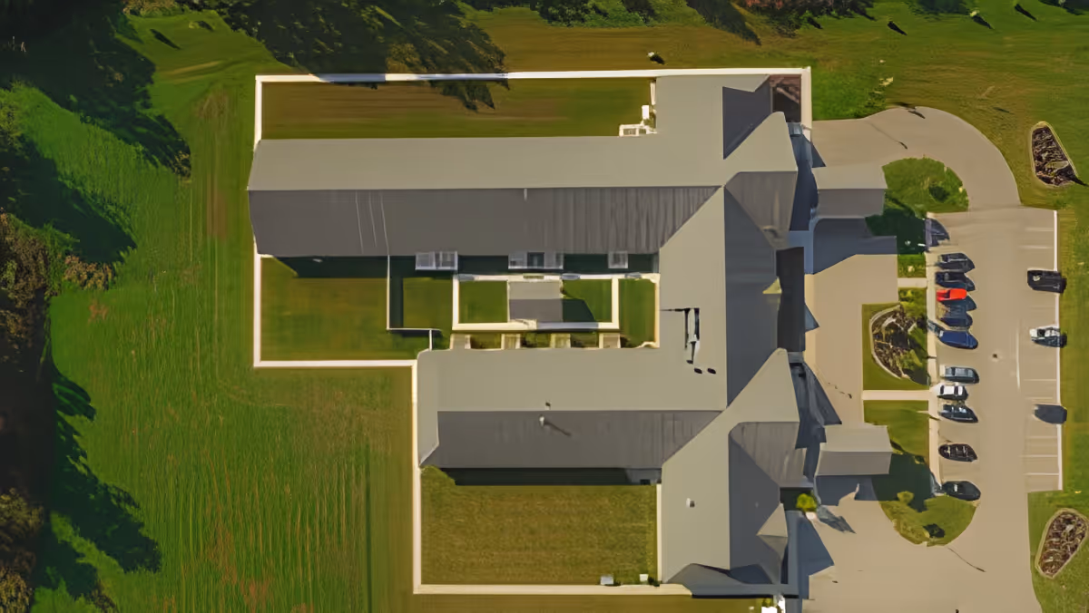 Aerial view of a senior living facility building with a gray roof, surrounded by green lawns and a parking lot with several cars parked. The building has a rectangular layout with an inner courtyard area.