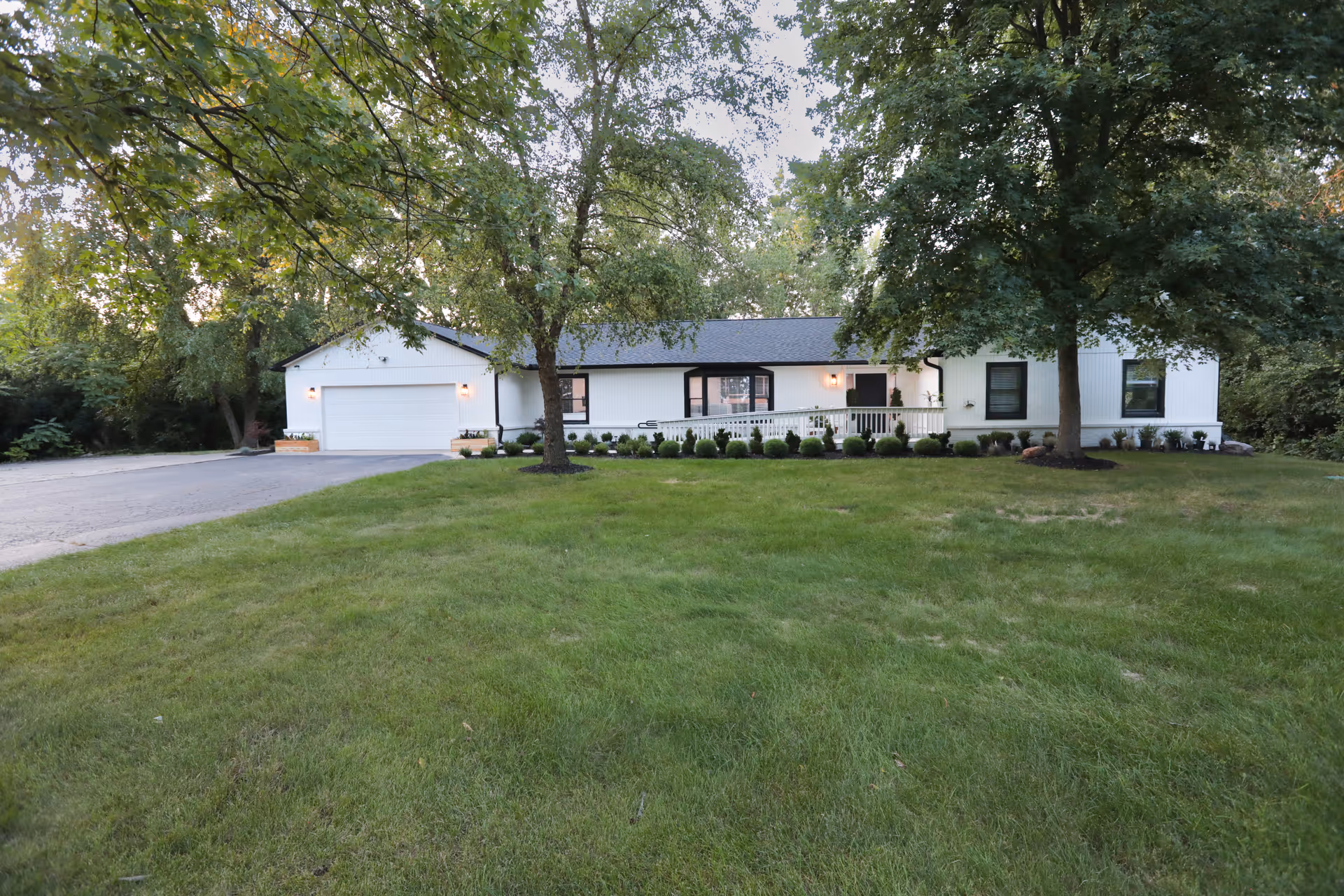 A single-story white building with a black roof surrounded by green grass and trees. The building has a garage on the left side, a ramp leading to the entrance, and small shrubs planted along the front. The scene is outdoors with natural lighting.