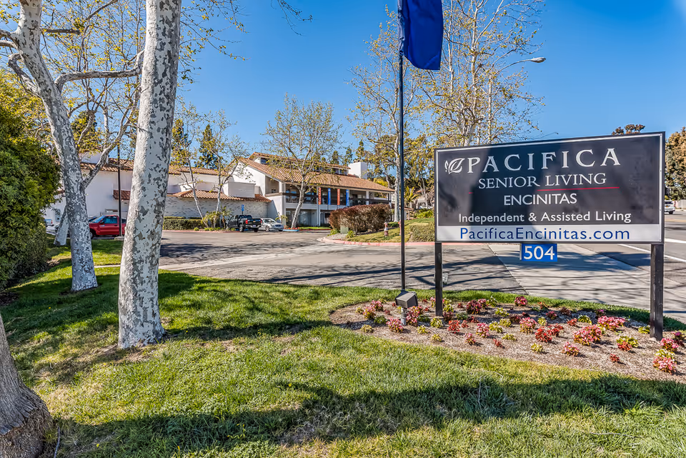 Outdoor view of Pacifica Senior Living Encinitas facility sign with building and parking lot in the background, surrounded by trees and grass under a clear blue sky.