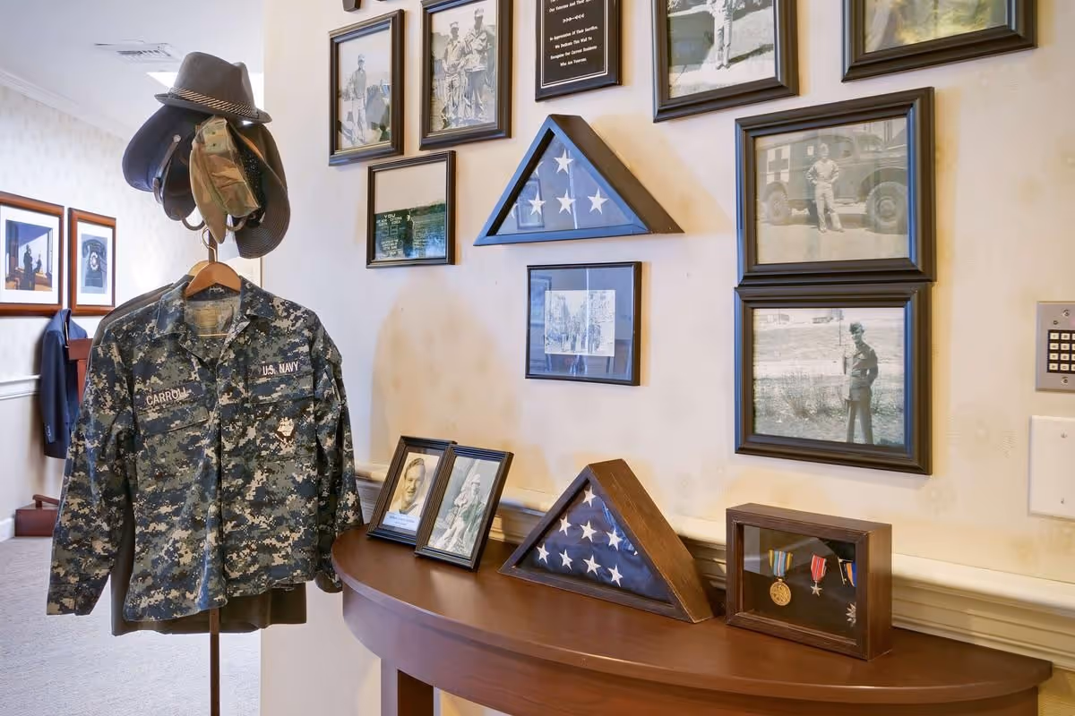 A hallway corner in a senior living facility decorated with military memorabilia including a U.S. Navy camouflage jacket on a coat rack with hats, framed black and white photographs on the wall, two folded American flags in triangular display cases on a wooden table, and a display case with military medals.