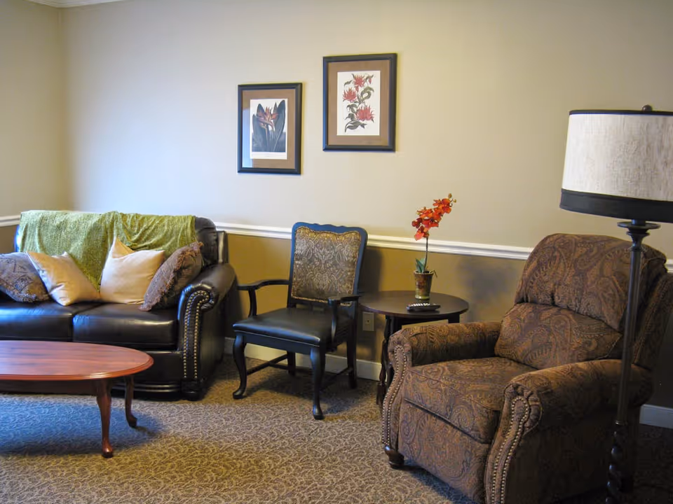A cozy living room area with a dark leather sofa adorned with multiple pillows and a green throw blanket, a patterned armchair, a wooden chair with a cushioned seat, a round side table with a small potted plant and a remote control, a wooden coffee table, and a floor lamp. Two framed floral artworks hang on the beige wall above the furniture.