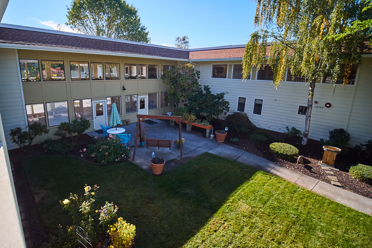 Sunlit courtyard with a grassy lawn, patio seating and a wooden swing surrounded by a two-story senior living building.