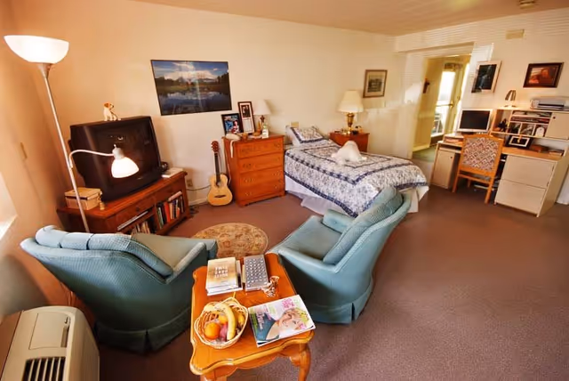 A cozy assisted living room featuring a single bed with a blue and white quilt, two green armchairs facing a small wooden table with a fruit basket and magazines, a wooden dresser with framed photos and a lamp, a guitar leaning against the wall, a TV on a wooden stand, and a desk with a computer and chair in the background.