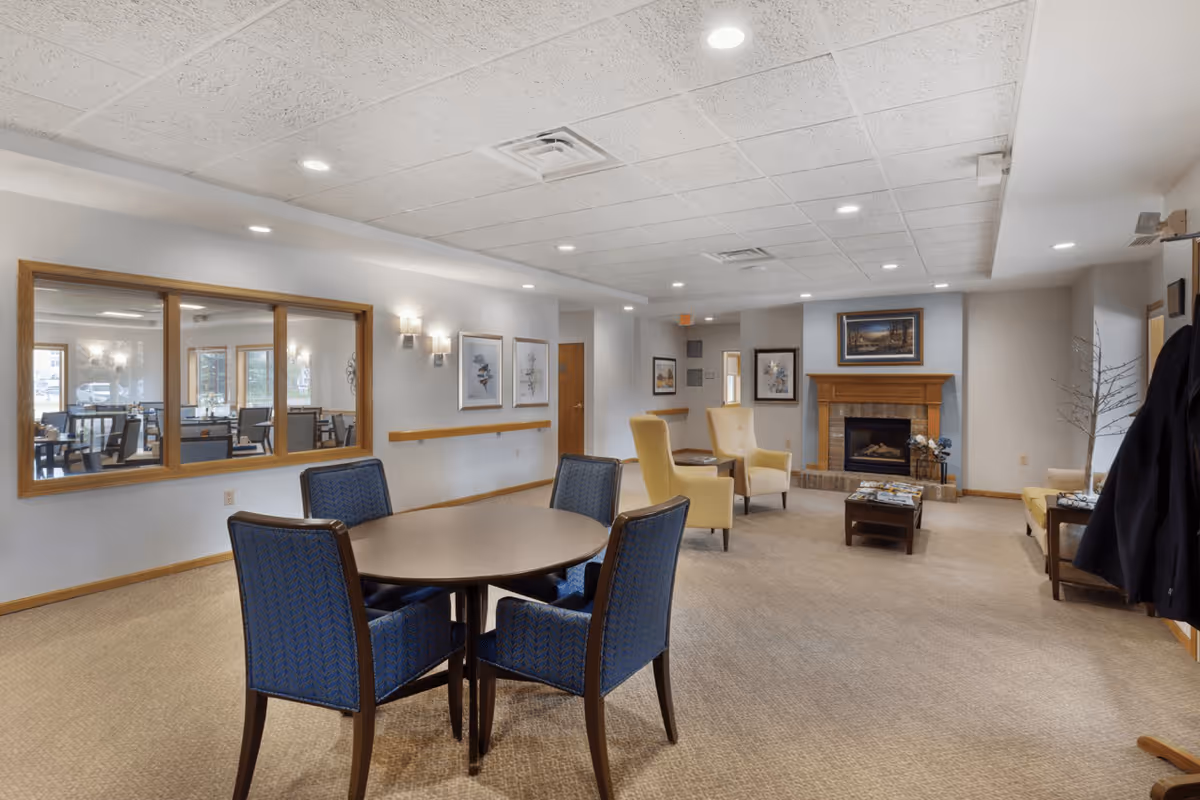 A senior living facility common area with a round table and four blue upholstered chairs in the foreground. The room features beige carpeting, a fireplace with a wooden mantel, two yellow armchairs, framed artwork on the walls, and a window looking into a dining area with tables and chairs.