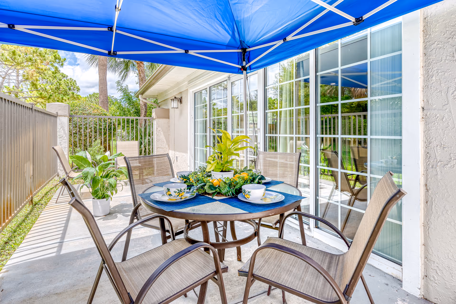 Outdoor patio area with a round glass table set for four with decorative plates, cups, and a centerpiece of green plants and lemons. The patio is shaded by a blue canopy and surrounded by a metal fence. There are additional potted plants and sliding glass doors leading inside the building.