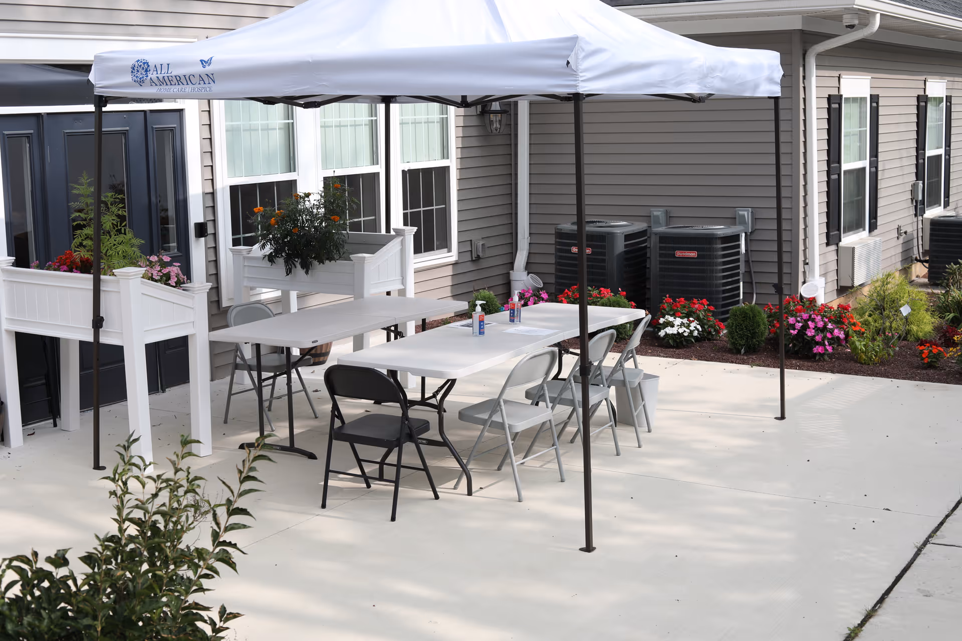 Outdoor patio area with a white canopy tent covering two folding tables and several folding chairs. There are flower planters with colorful flowers near the building's windows and air conditioning units. The building has beige siding and multiple windows.
