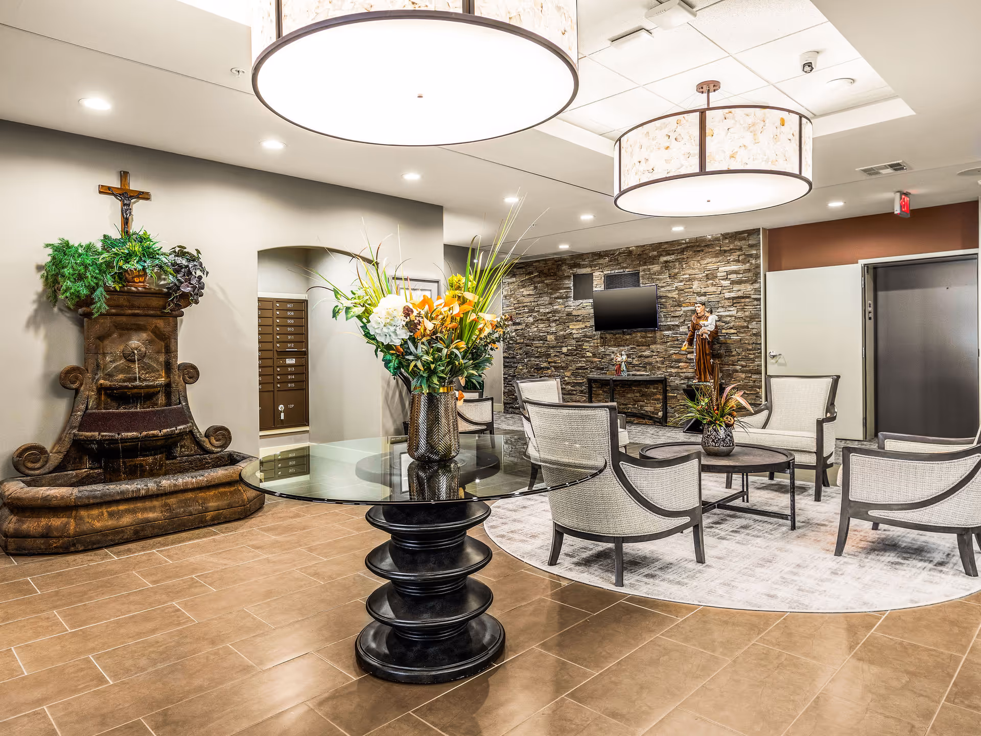 A well-lit senior living facility common area with a round glass table holding a floral arrangement in the foreground. There are several cushioned chairs arranged around a circular rug, a stone accent wall with a mounted TV, and a statue. A decorative water fountain with plants and a crucifix is on the left side of the room.