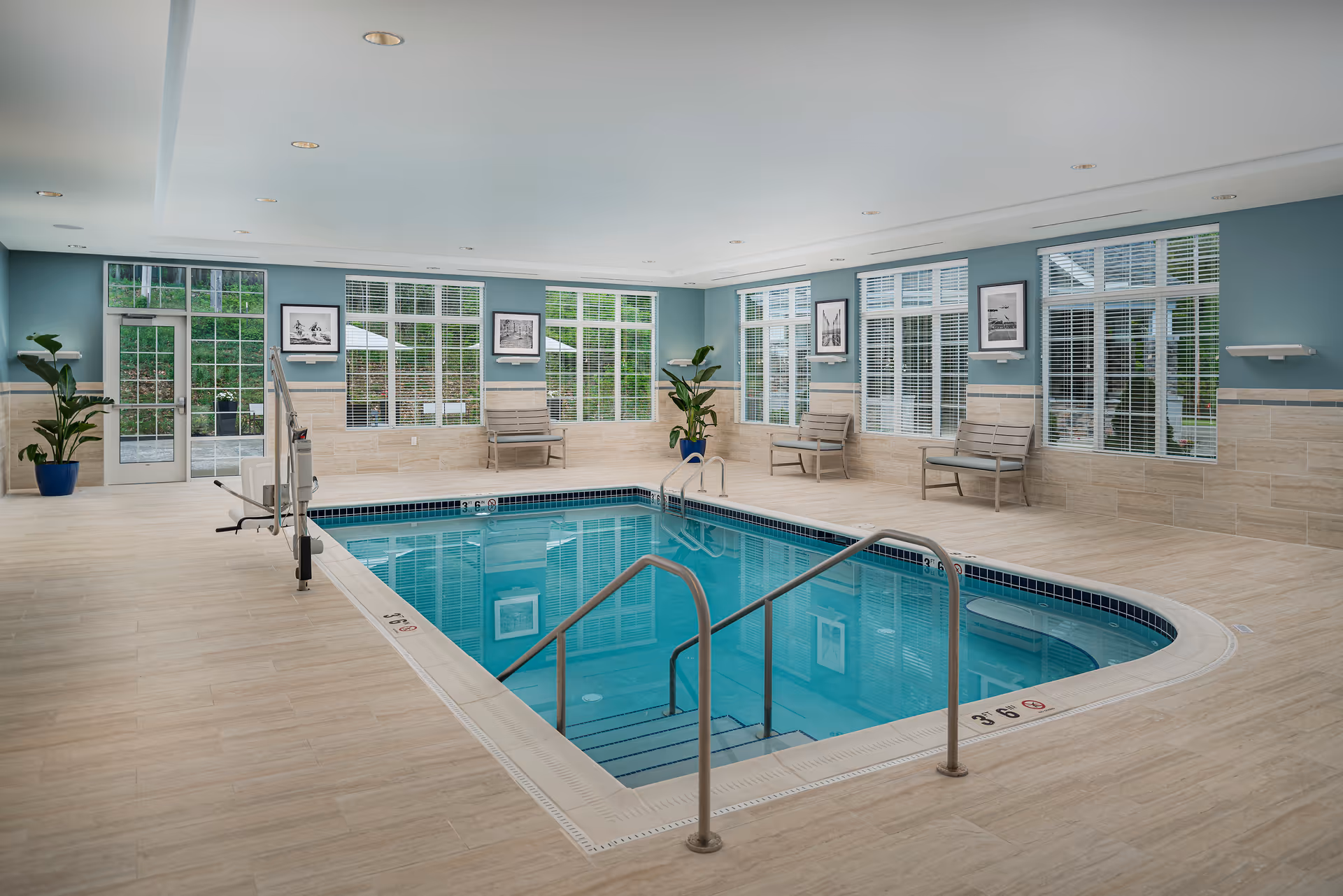 Indoor swimming pool area with clear blue water, surrounded by beige tiled floor and light blue walls. Several large windows with white blinds allow natural light to enter. There are a few benches and potted plants along the walls, and a pool lift for accessibility is installed at one corner of the pool.