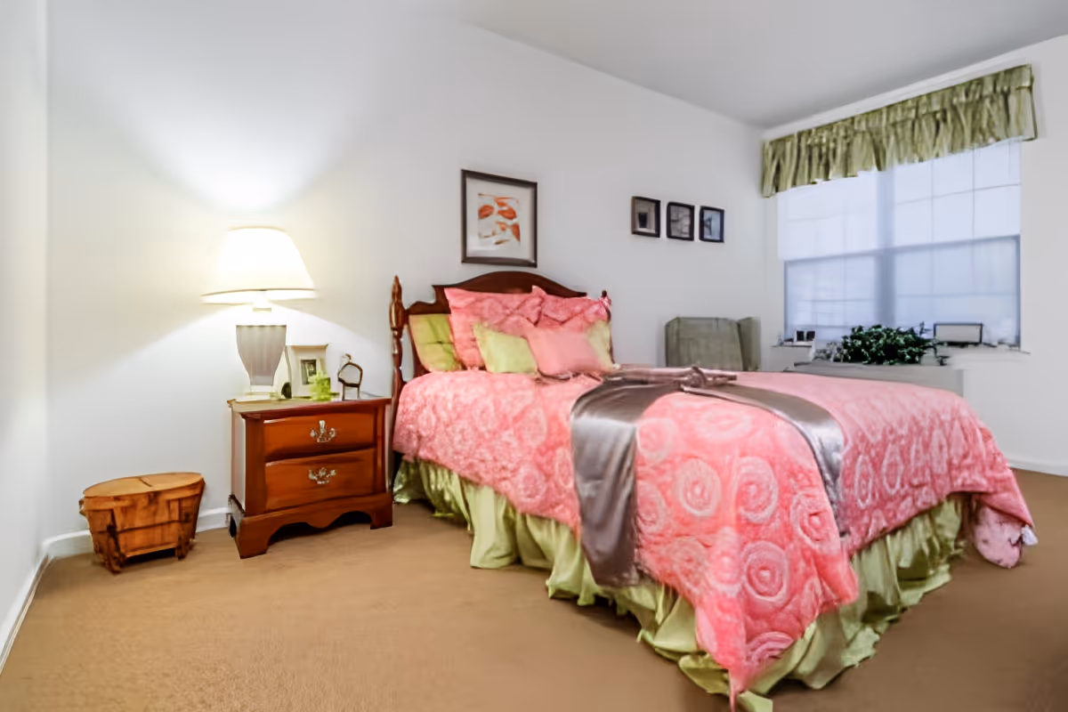 A bedroom with a bed covered in pink and green bedding, including multiple pillows and a pink blanket with a circular pattern. Next to the bed is a wooden nightstand with a lamp, picture frame, and small decorative items. There is a small wooden basket on the floor beside the nightstand. The room has beige carpet, white walls, and a window with green valance curtains and white blinds. A green armchair is positioned near the window.