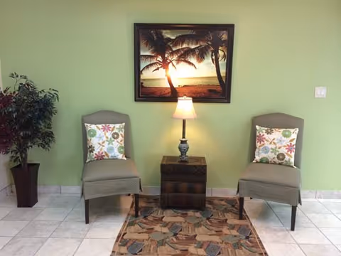 Two cushioned chairs with floral pillows are placed on either side of a small wooden table with a lamp on it. A framed picture of a sunset with palm trees hangs on a light green wall behind the chairs. A potted plant is positioned to the left of the chairs, and a patterned rug lies on the tiled floor in front of them.