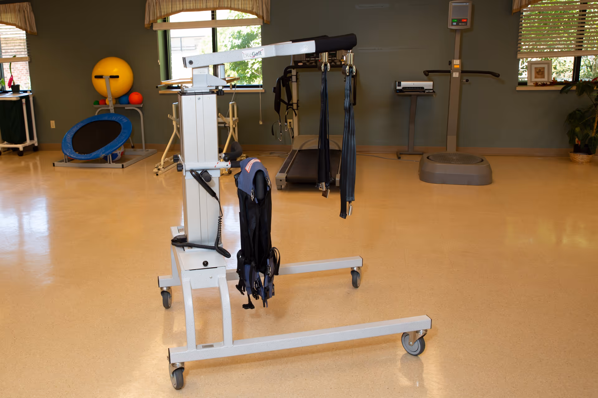 Physical therapy room with a mobile patient lift in the foreground, treadmill, exercise equipment and therapy balls in the background.