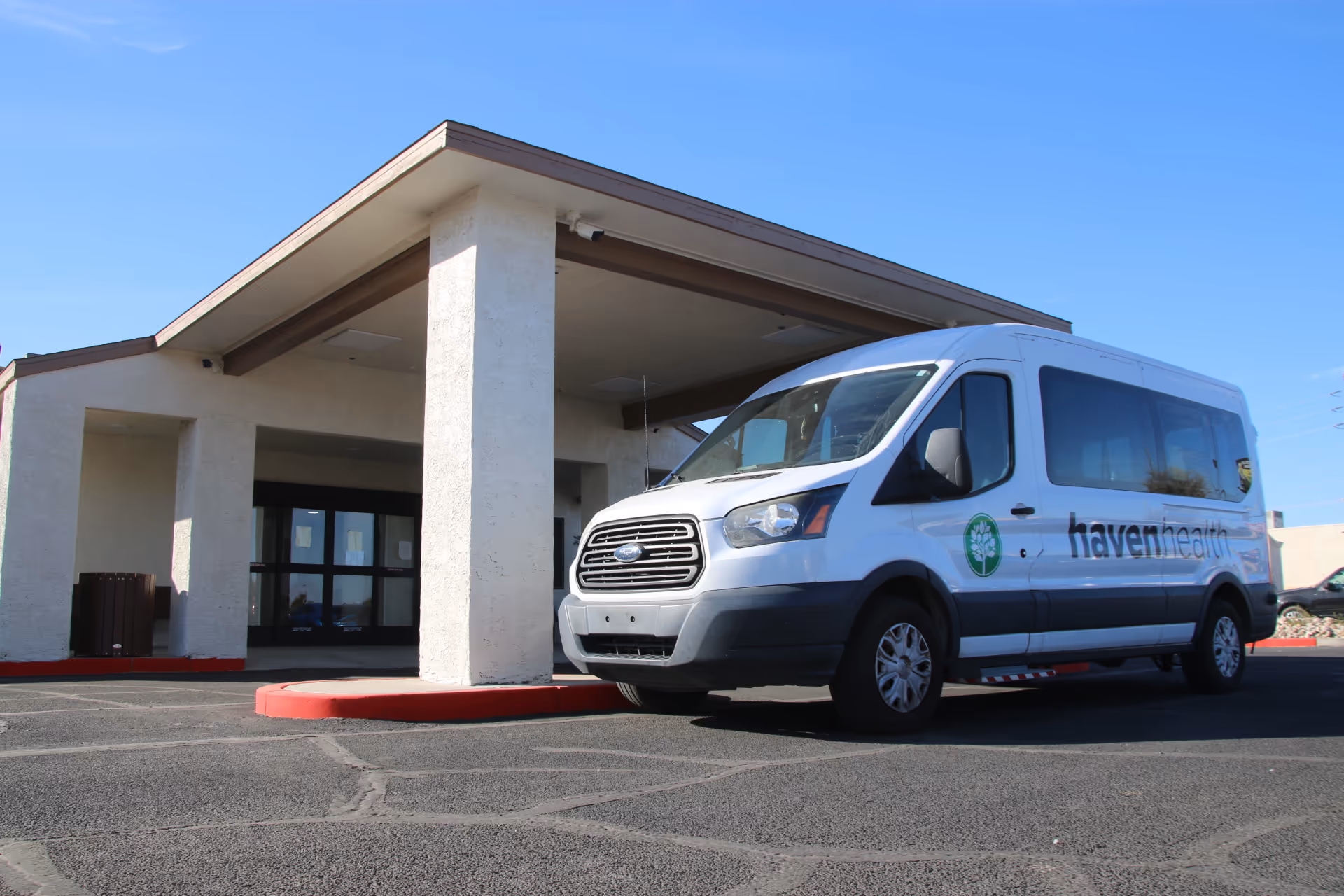 A white shuttle van with the logo and text 'haven health' parked in front of the entrance to a building under a covered driveway on a clear sunny day.