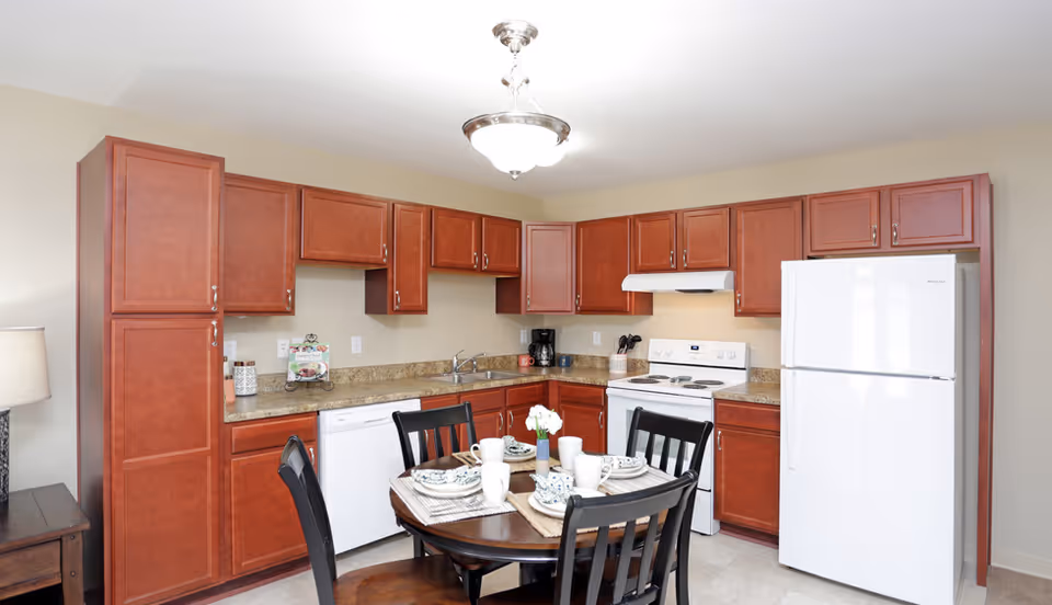 Well-lit kitchen with cherry wood cabinets, white appliances and a small round dining table set for four.