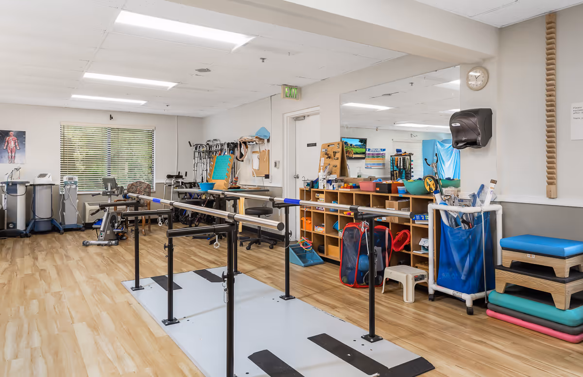 Physical therapy room with parallel bars for walking exercises, various exercise equipment, chairs, and storage shelves filled with therapy supplies. The room has wood flooring, a large mirror on one wall, and a window with blinds.