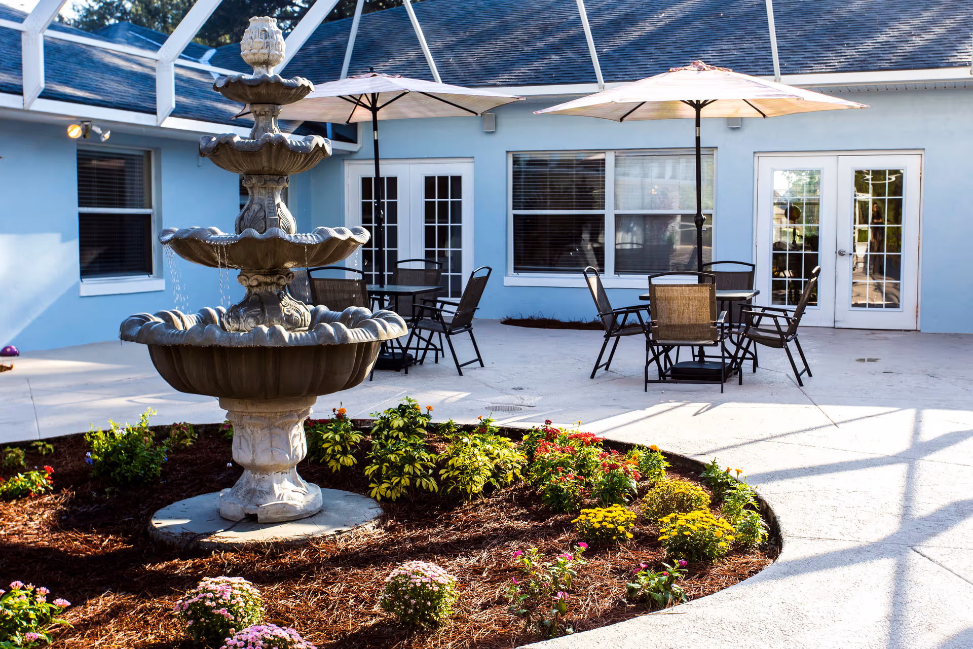 Outdoor patio area with a three-tiered stone fountain surrounded by a flower bed with various plants and flowers. There are two tables with umbrellas and several chairs on a concrete surface, adjacent to a light blue building with windows and glass doors.
