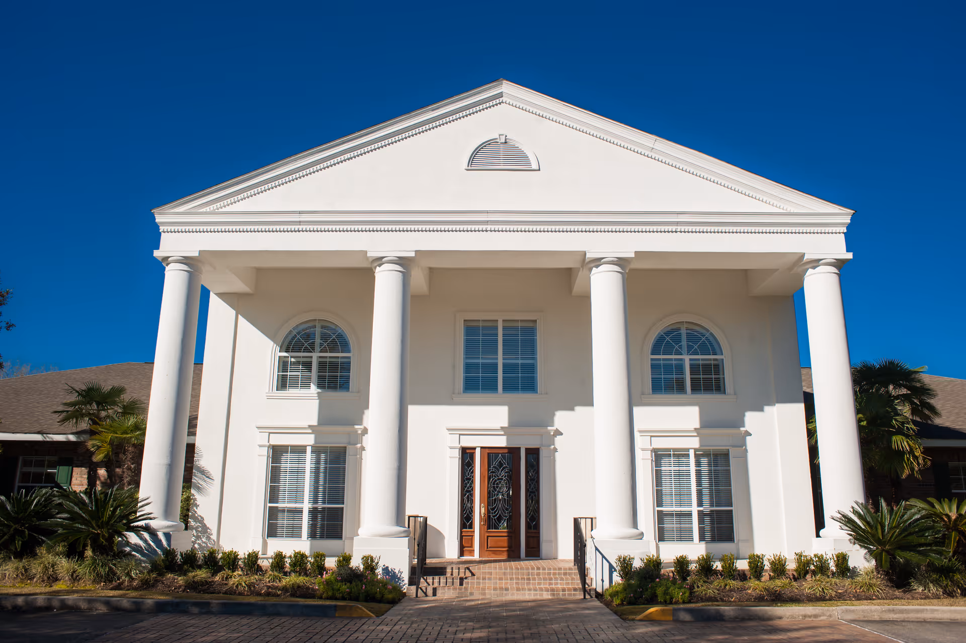 Front exterior view of a large white building with classical architecture featuring four tall columns, arched windows, and a wooden double door entrance under a clear blue sky.