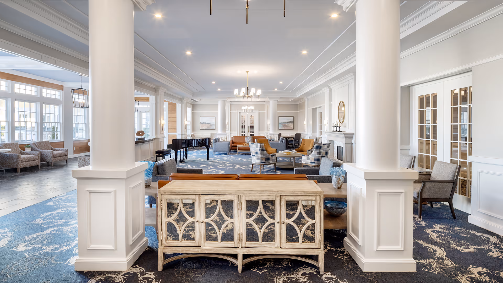 A spacious and elegant living room area in a senior living facility with white walls, large white columns, and a blue patterned carpet. The room features multiple seating arrangements including sofas, armchairs, and a round table with chairs. A black grand piano is positioned near the windows, and there are chandeliers hanging from the ceiling. Large windows allow natural light to fill the space.