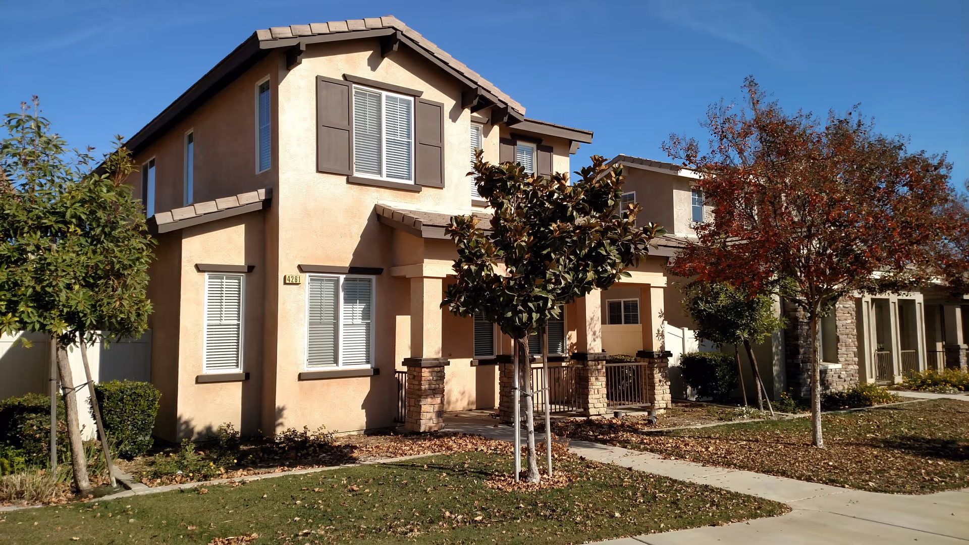 Exterior view of a two-story residential building with beige stucco walls, brown roof tiles, and several windows with shutters. There are small trees and bushes in the front yard, a concrete walkway leading to the entrance, and fallen leaves scattered on the grass and sidewalk under a clear blue sky.