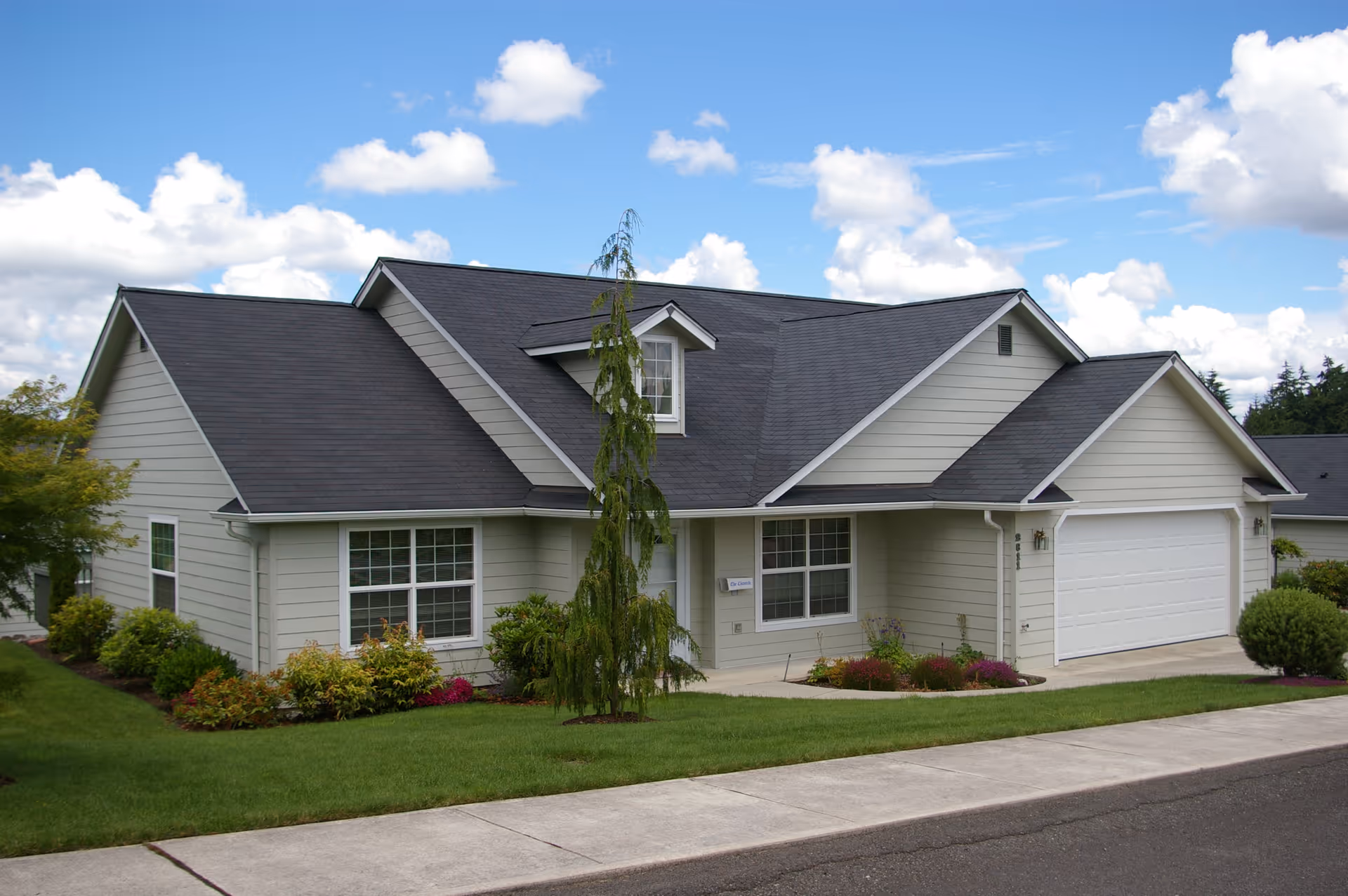 Exterior view of a single-story residential building with light gray siding, a dark gray roof, a two-car garage, and a well-maintained lawn with shrubs and a small tree in front under a partly cloudy sky.