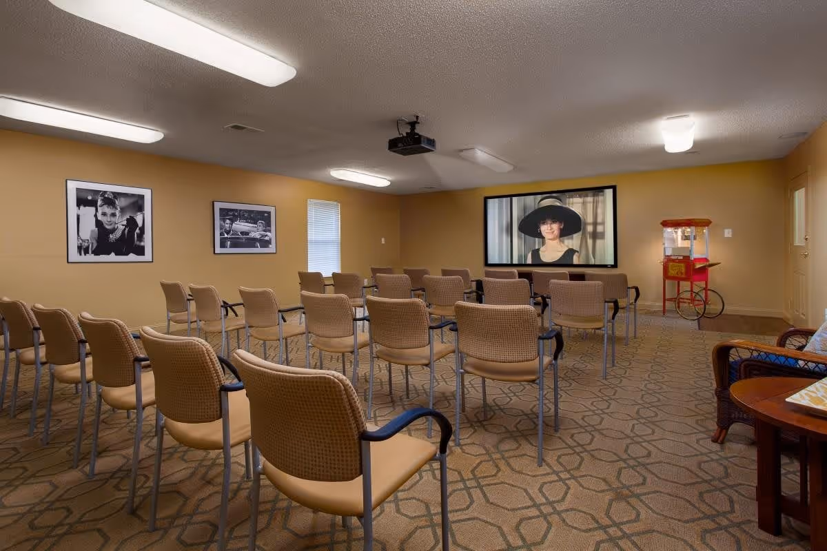 Small community media/theater room with rows of chairs facing a projection screen showing a woman, framed photos on the walls and a popcorn machine at the side.