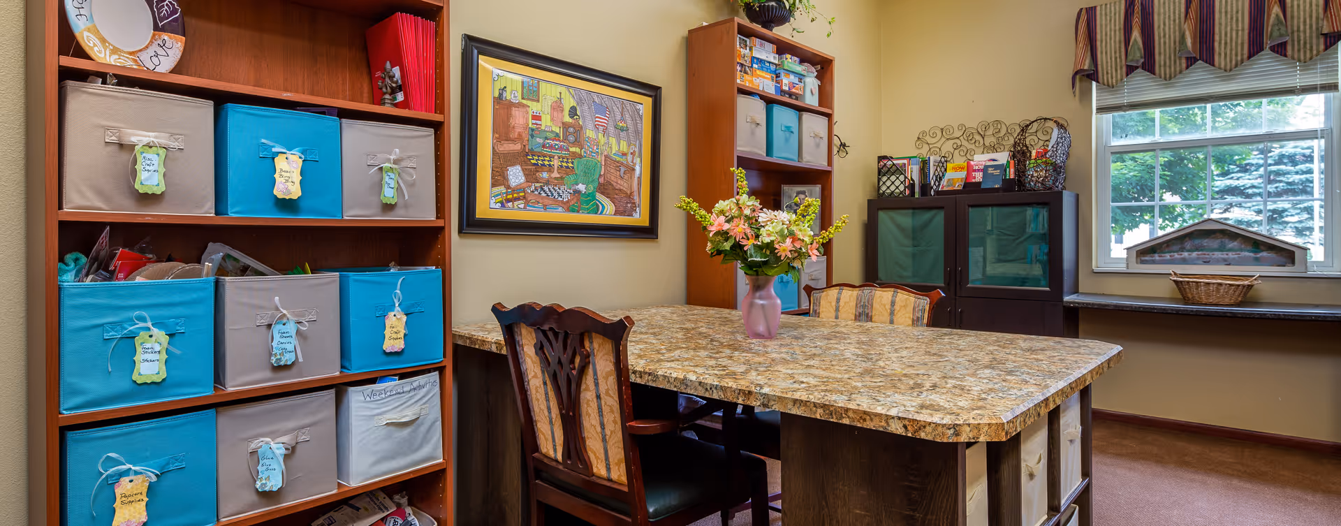 A well-organized room with a large table in the center, surrounded by chairs. On the left side, there is a wooden shelving unit with colorful storage bins labeled with tags. A framed picture hangs on the wall above the table. In the background, there is another shelving unit with more storage bins and a window with a striped valance, showing greenery outside. A vase with flowers is placed on the table.