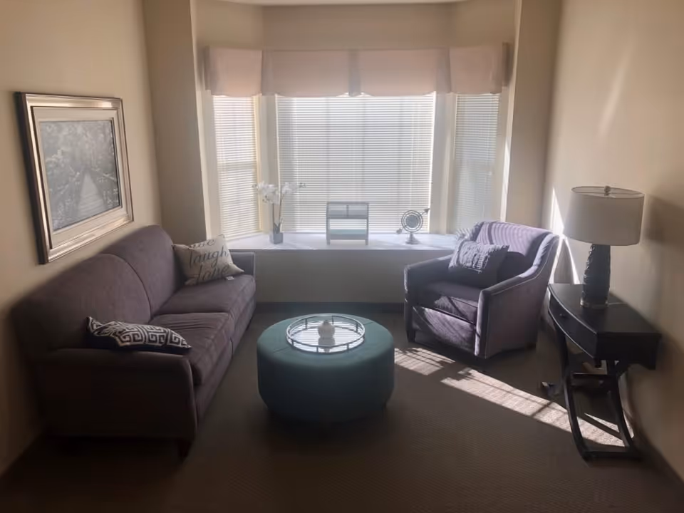 A cozy living room with a purple sofa and matching armchair, a round teal ottoman with a decorative tray, a side table with a lamp, and a large window with blinds and a valance letting in natural light. A framed picture hangs on the wall above the sofa.
