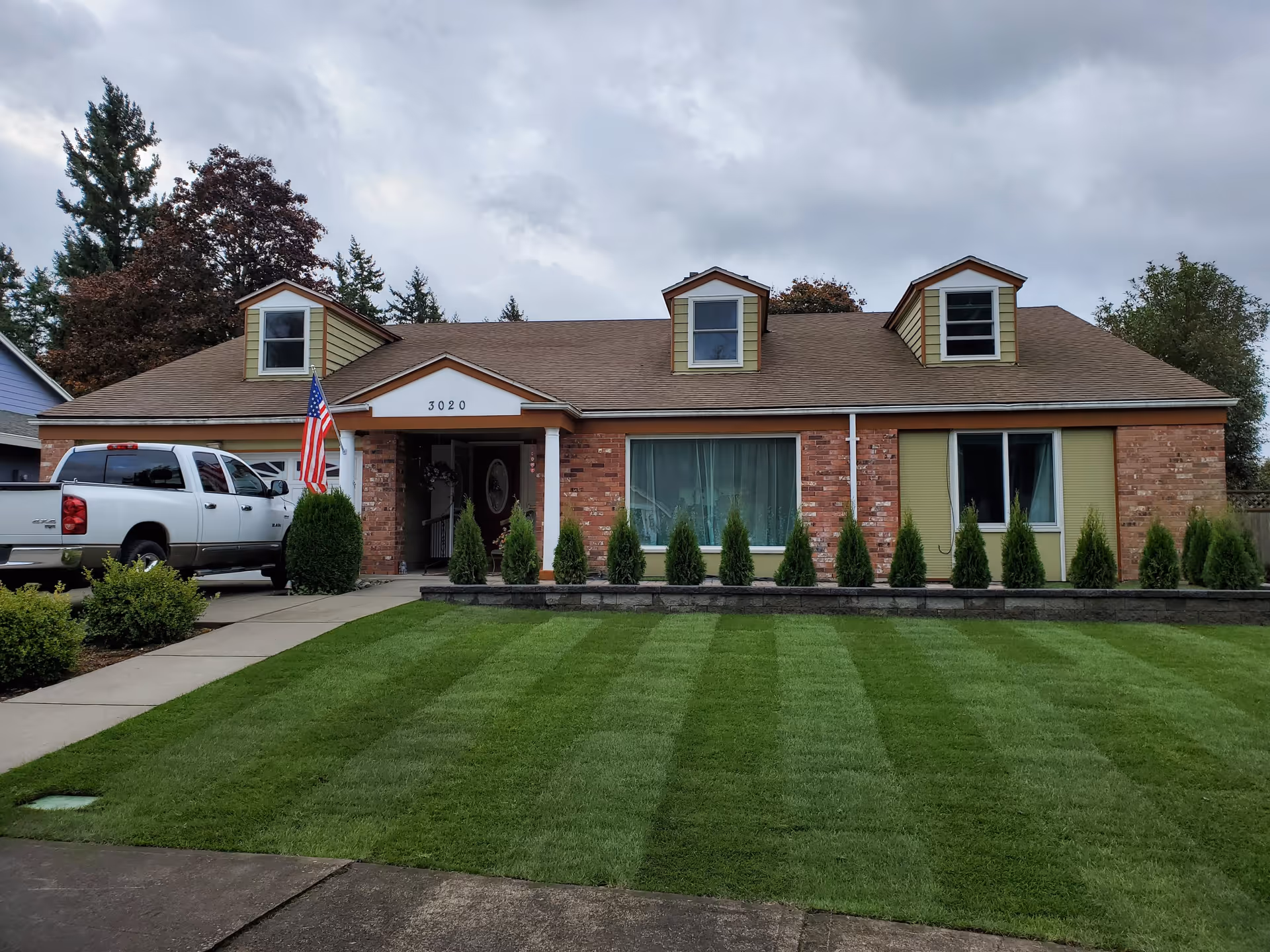 Front exterior view of a single-story brick house with three dormer windows on the roof, a well-maintained green lawn with striped mowing patterns, small shrubs along the front, an American flag near the entrance, and a white pickup truck parked in the driveway.