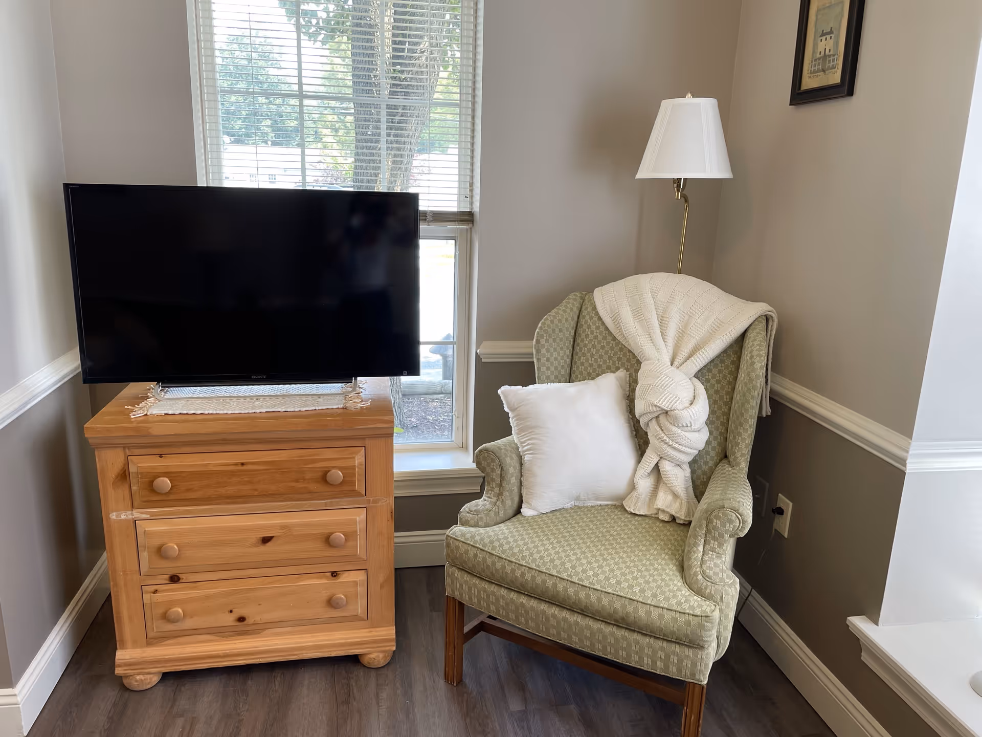 A cozy corner of a room featuring a green upholstered armchair with a white pillow and a white knitted blanket draped over the back. Next to the chair is a wooden three-drawer dresser with a flat-screen TV on top. A window with blinds is behind the dresser, and a floor lamp with a white shade stands beside the chair. A small framed picture hangs on the wall above the lamp.