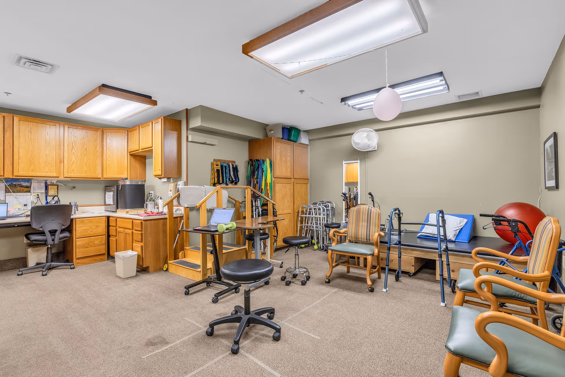 A therapy or rehabilitation room with wooden cabinets and a small desk area on the left. The room contains various physical therapy equipment including parallel bars, walkers, exercise balls, and chairs arranged around the space. The walls are light beige and the floor is carpeted.