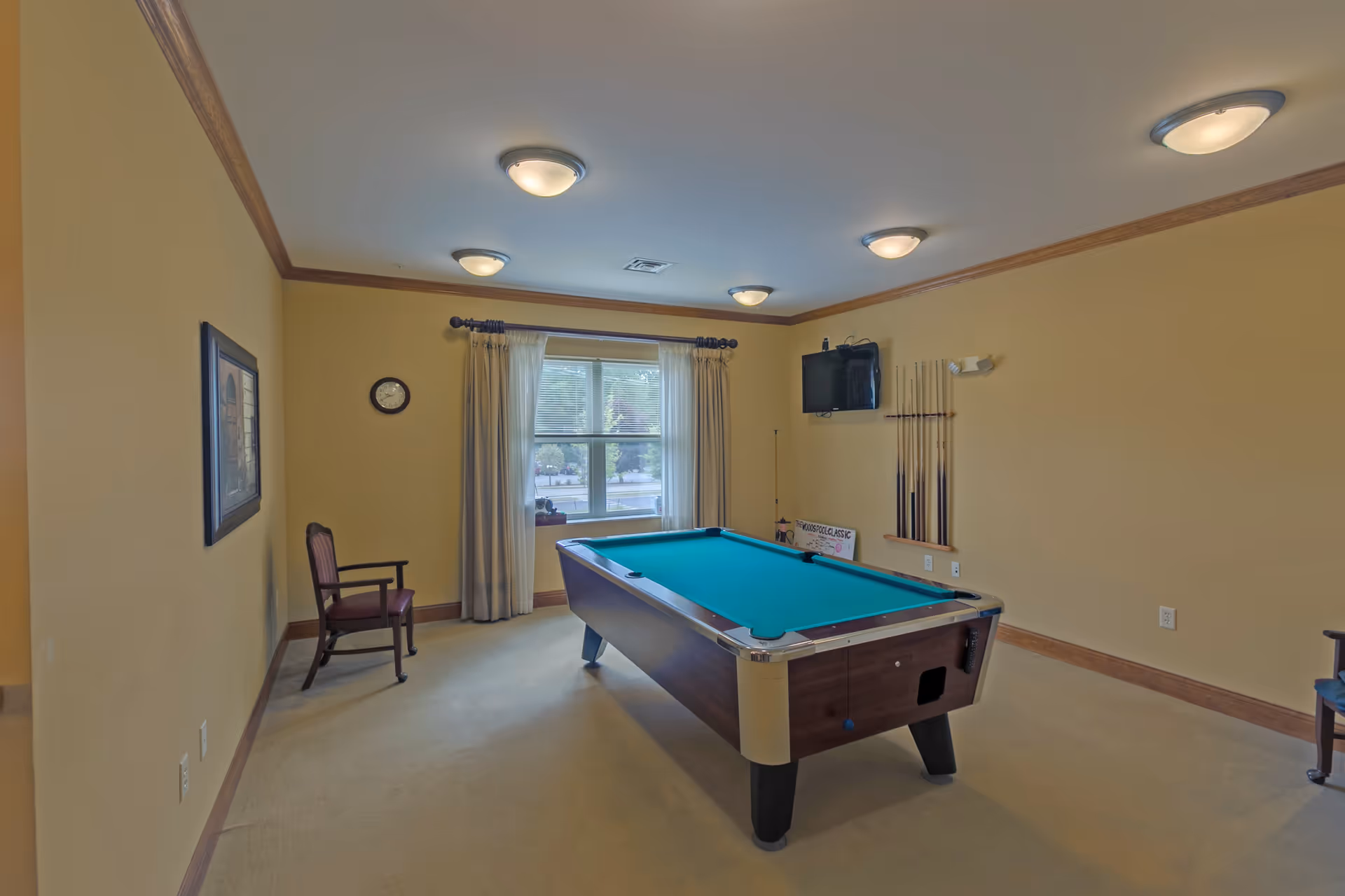 A recreation room with a pool table in the center, a wall-mounted TV, pool cues on a rack, a window with curtains, a clock on the wall, and a wooden chair with armrests.