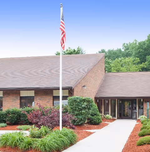 Brick single-story building entrance with an American flag, a concrete walkway, and landscaped shrubs.