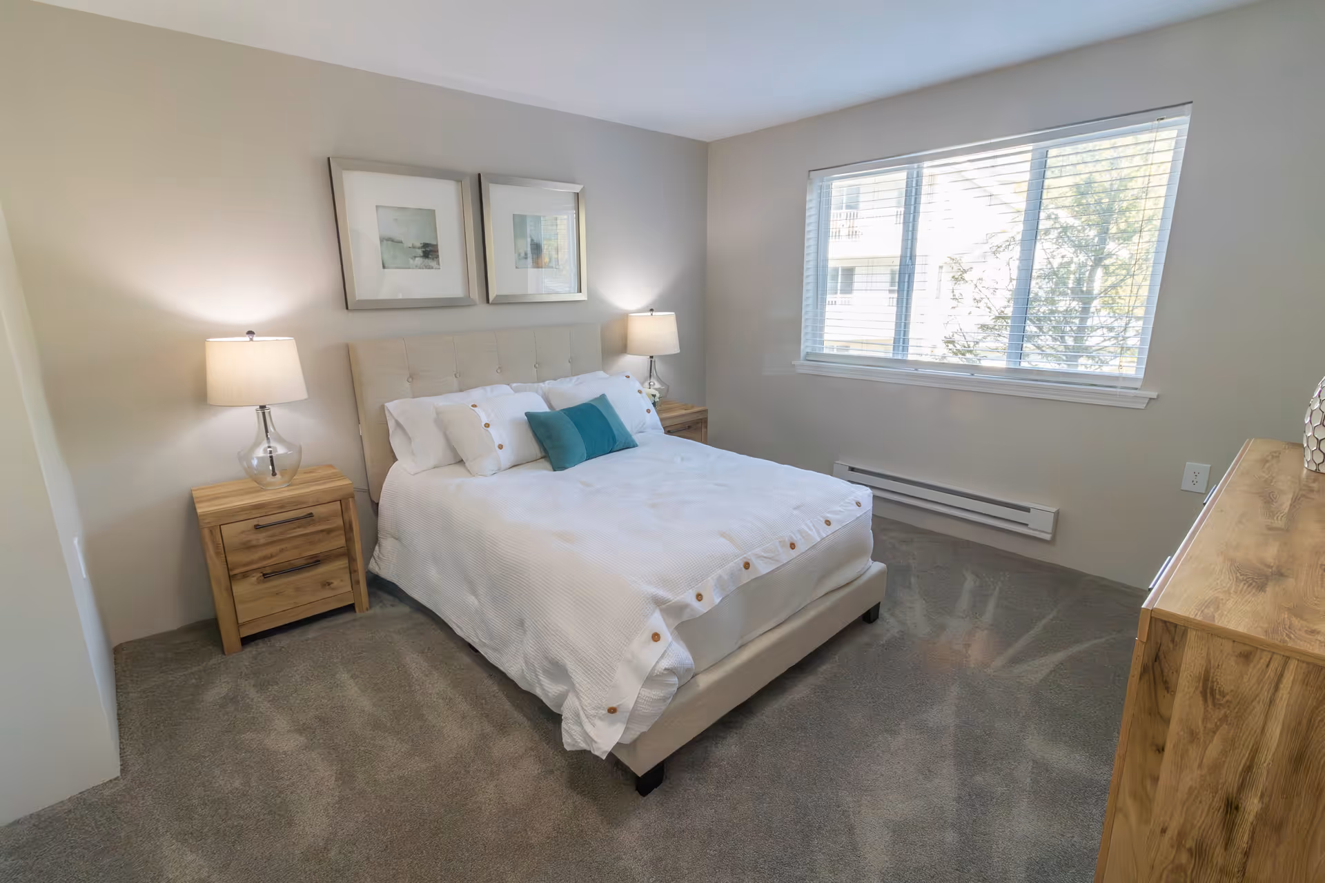 A bright bedroom with a large window, a bed with white bedding and blue accent pillows, two wooden nightstands with lamps, two framed pictures on the wall above the bed, and a wooden dresser on the right side.