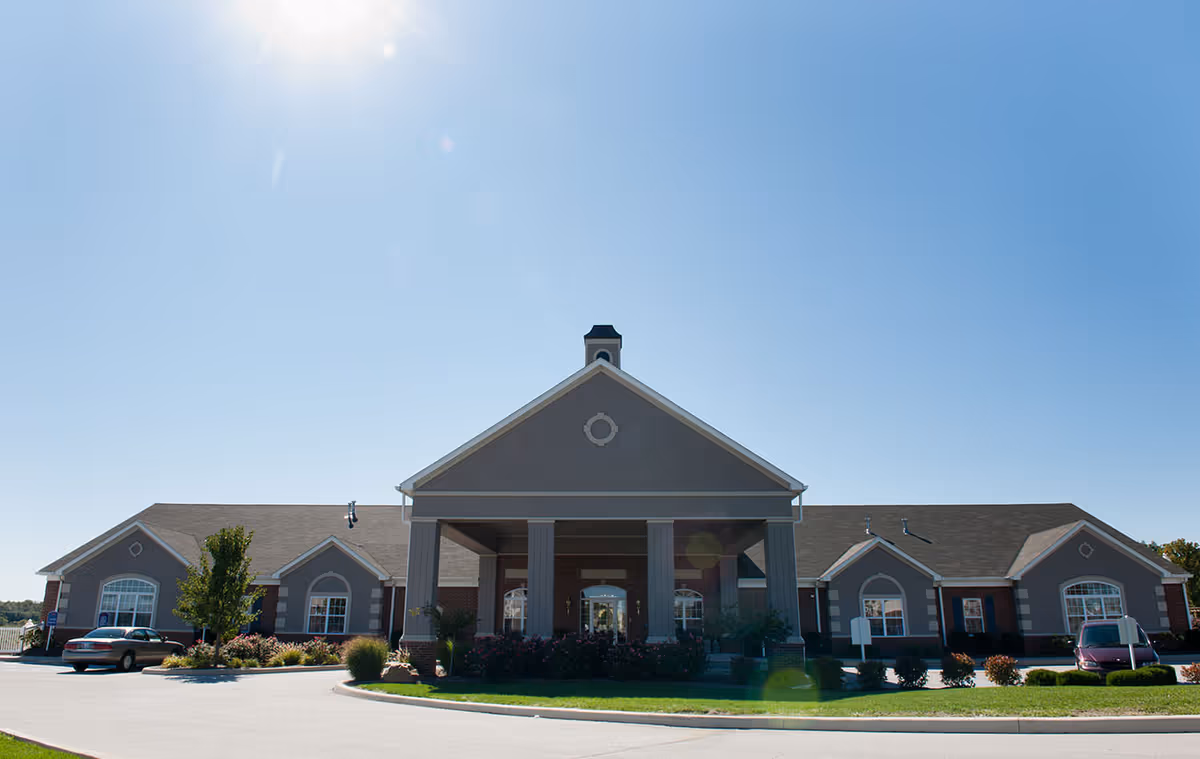 Front exterior of a single-story assisted living building with a central covered entrance, columns, landscaping, and parked cars.