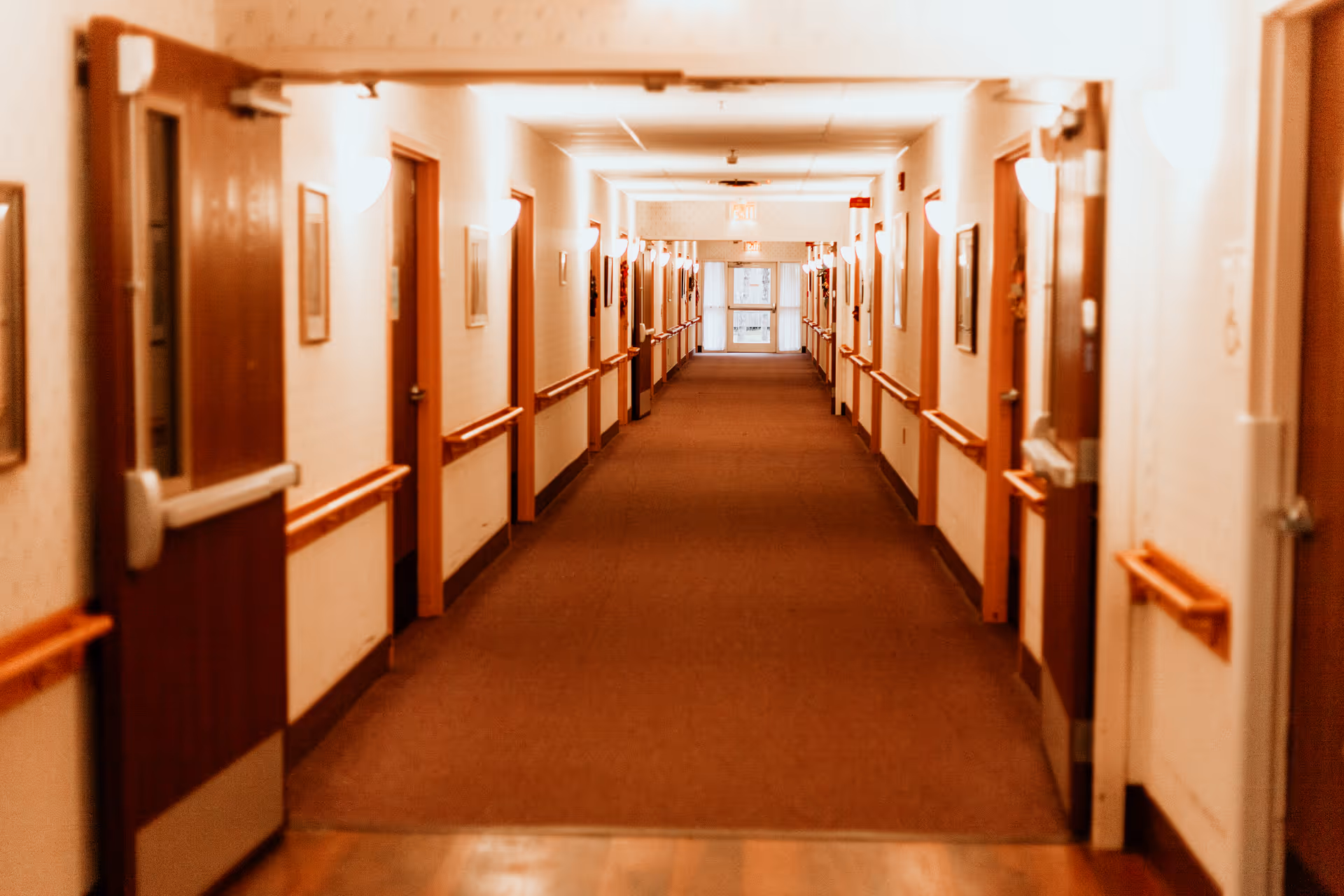 A long, well-lit hallway in a senior living facility with multiple closed wooden doors on both sides, handrails along the walls, and framed pictures hanging above the handrails.