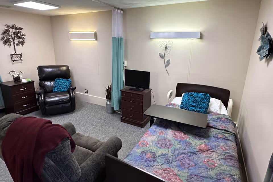 Assisted-living room with a single bed covered in a floral quilt, reclining chairs, a TV on a dresser, and wall decorations.