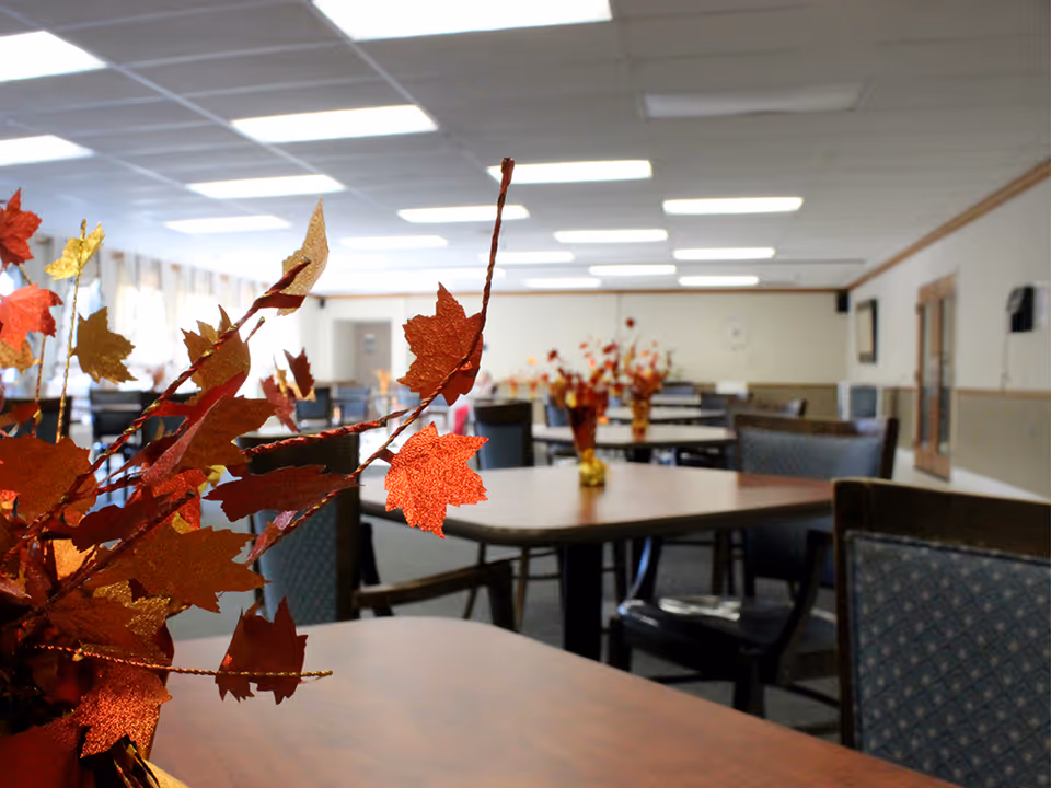 Interior view of a dining room with multiple tables and chairs arranged neatly. Each table has a centerpiece with artificial autumn leaves in red, orange, and gold colors. The room has a drop ceiling with fluorescent lighting and windows along one side letting in natural light.