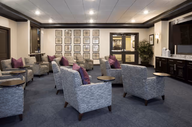 A spacious and well-lit lounge area with multiple gray upholstered armchairs arranged in pairs, each with a small round side table. The chairs have purple accent pillows. The room features a dark carpet with a subtle pattern, a wall decorated with a grid of framed artwork, and a large flat-screen TV mounted above a dark wood cabinet. There are double glass doors and a large potted plant near the entrance.