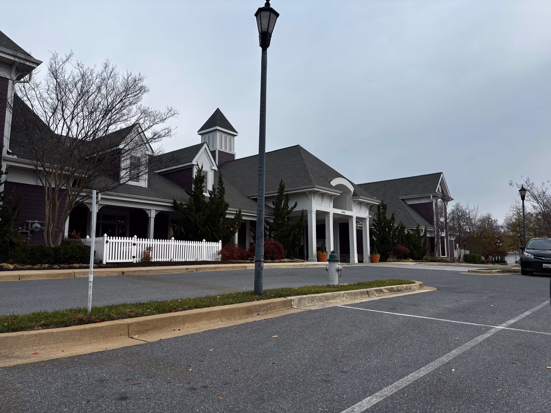 Front entrance and parking area of a large senior living building with white columns and a picket fence under an overcast sky.
