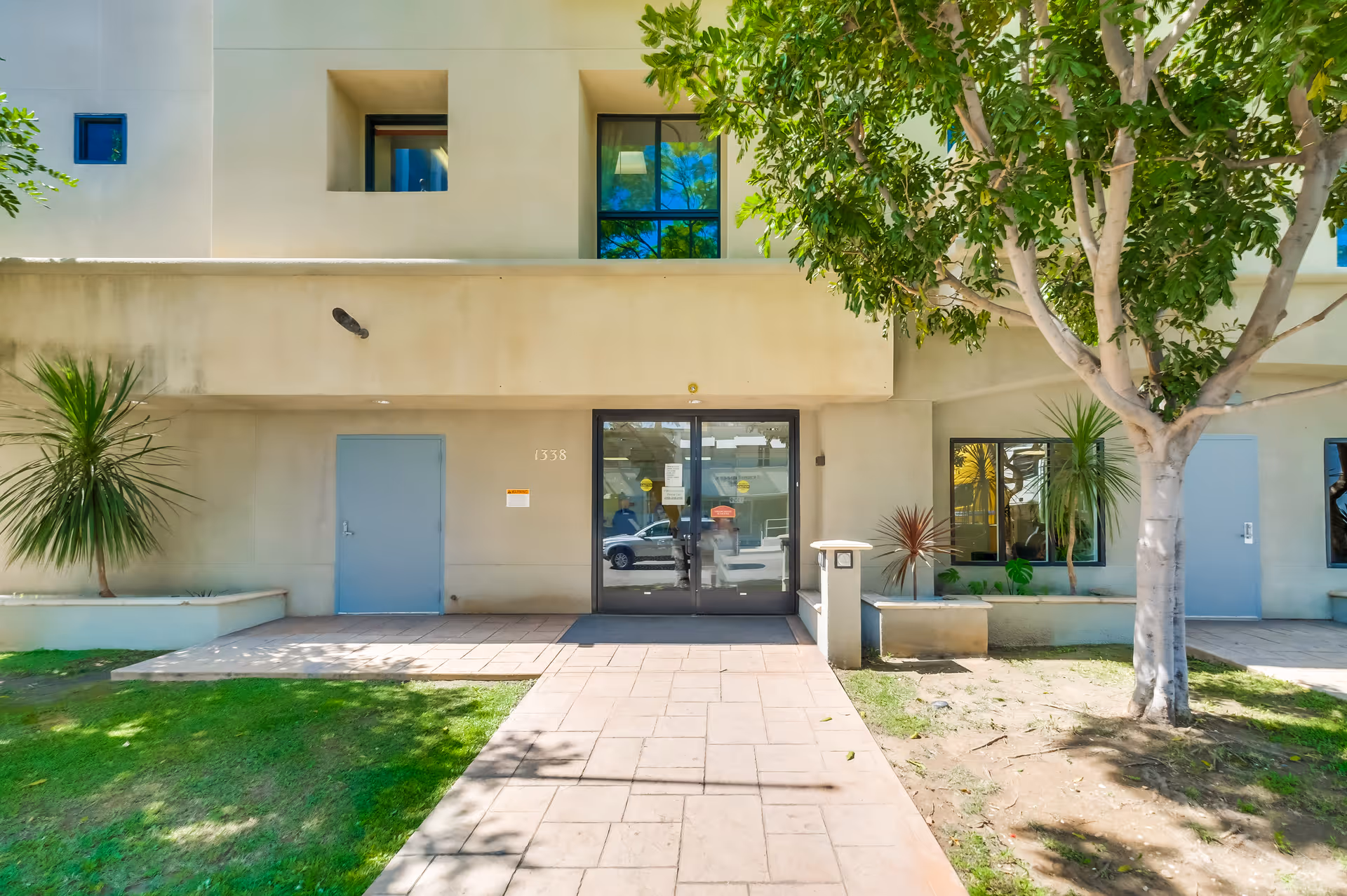 Exterior view of the entrance to Santa Monica Rehabilitation Center, featuring a beige building with glass double doors, two blue side doors, windows, and surrounding greenery including a tree and plants.