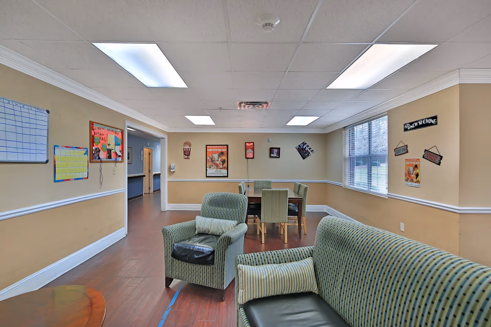 A common area in an assisted living facility with beige walls and wood flooring. The room features a striped armchair and sofa with cushions in the foreground, and a dining table with six chairs in the background. The walls are decorated with colorful posters, a whiteboard, and signs including one that says 'Now Showing'. Large windows with blinds allow natural light into the room.