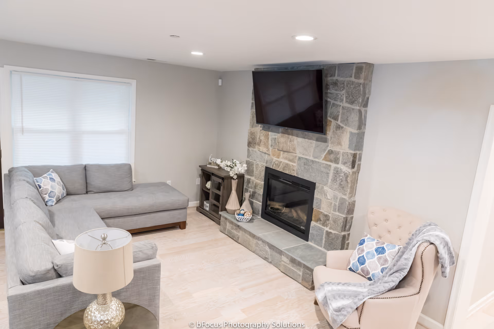 A cozy living room with a gray sectional sofa, a beige armchair with a patterned pillow and a gray throw blanket, a stone fireplace with a mounted flat-screen TV above it, a small wooden cabinet with decorative items, and a table lamp on a side table.