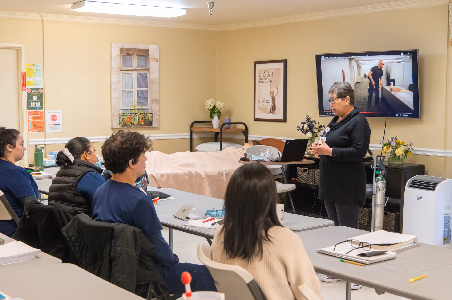 A group of staff sit at tables in a training room as an instructor speaks beside a TV and a hospital bed.
