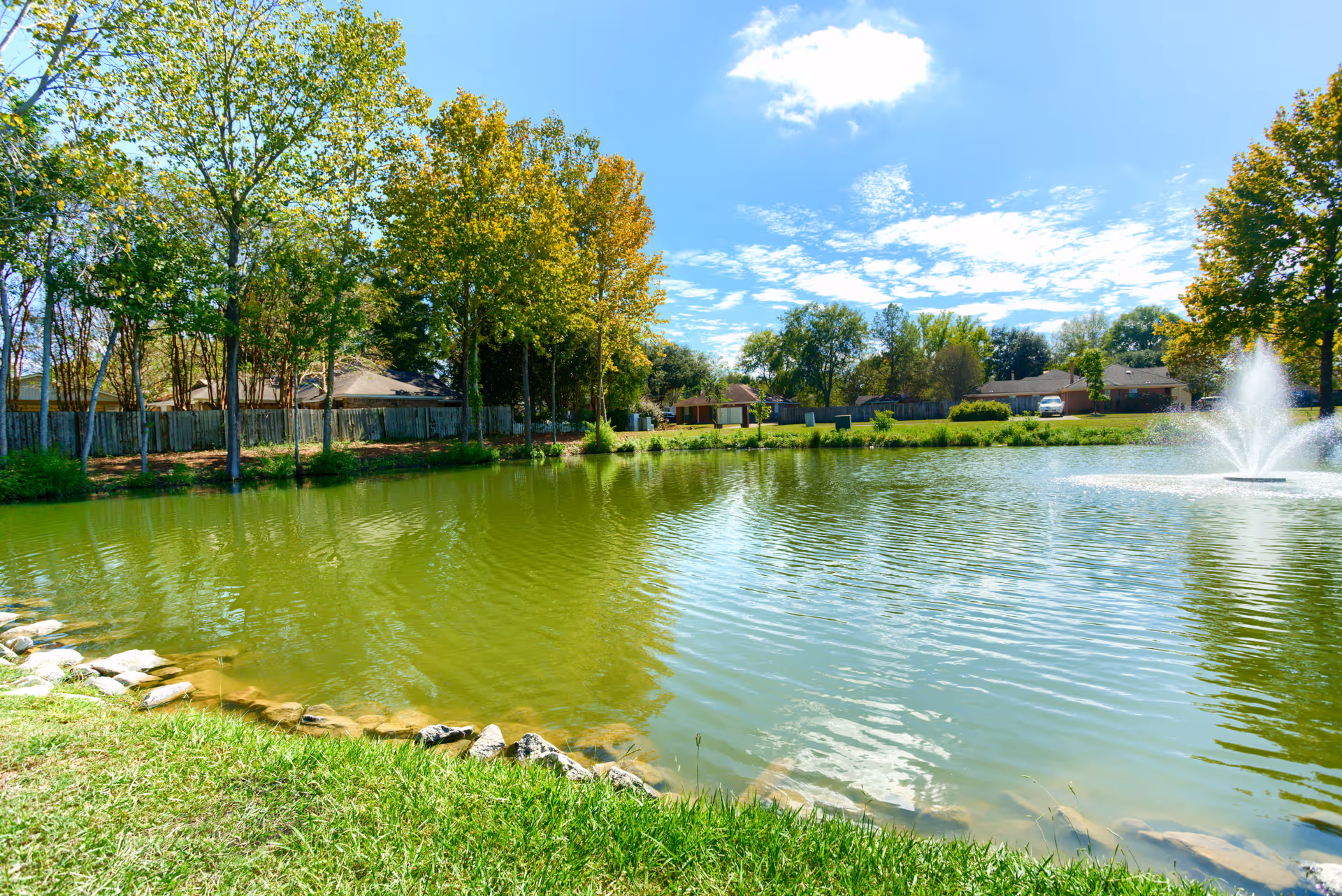A peaceful outdoor scene featuring a small pond with a water fountain in the middle, surrounded by green grass and trees. In the background, there are houses and a wooden fence under a partly cloudy blue sky.