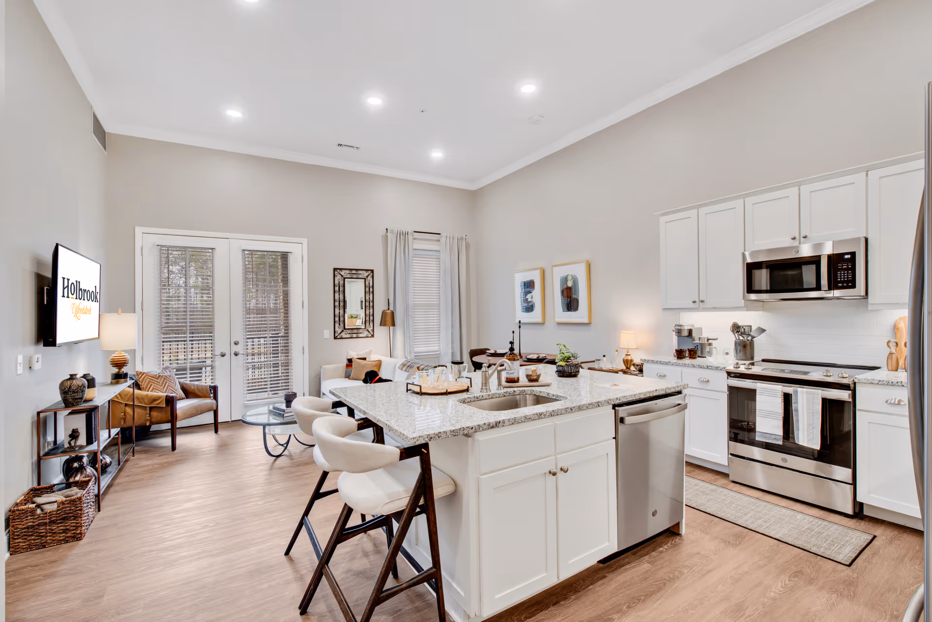 A bright and modern open-concept kitchen and living area in a senior living facility. The kitchen features white cabinets, stainless steel appliances including a microwave and stove, and a large granite island with a sink and three cushioned bar stools. The living area has a white sofa, a small table, a floor lamp, and a wall-mounted TV displaying the Holbrook Woodstock logo. French doors with blinds lead outside, and the room is decorated with framed artwork, a mirror, and various decorative items on shelves and tables.