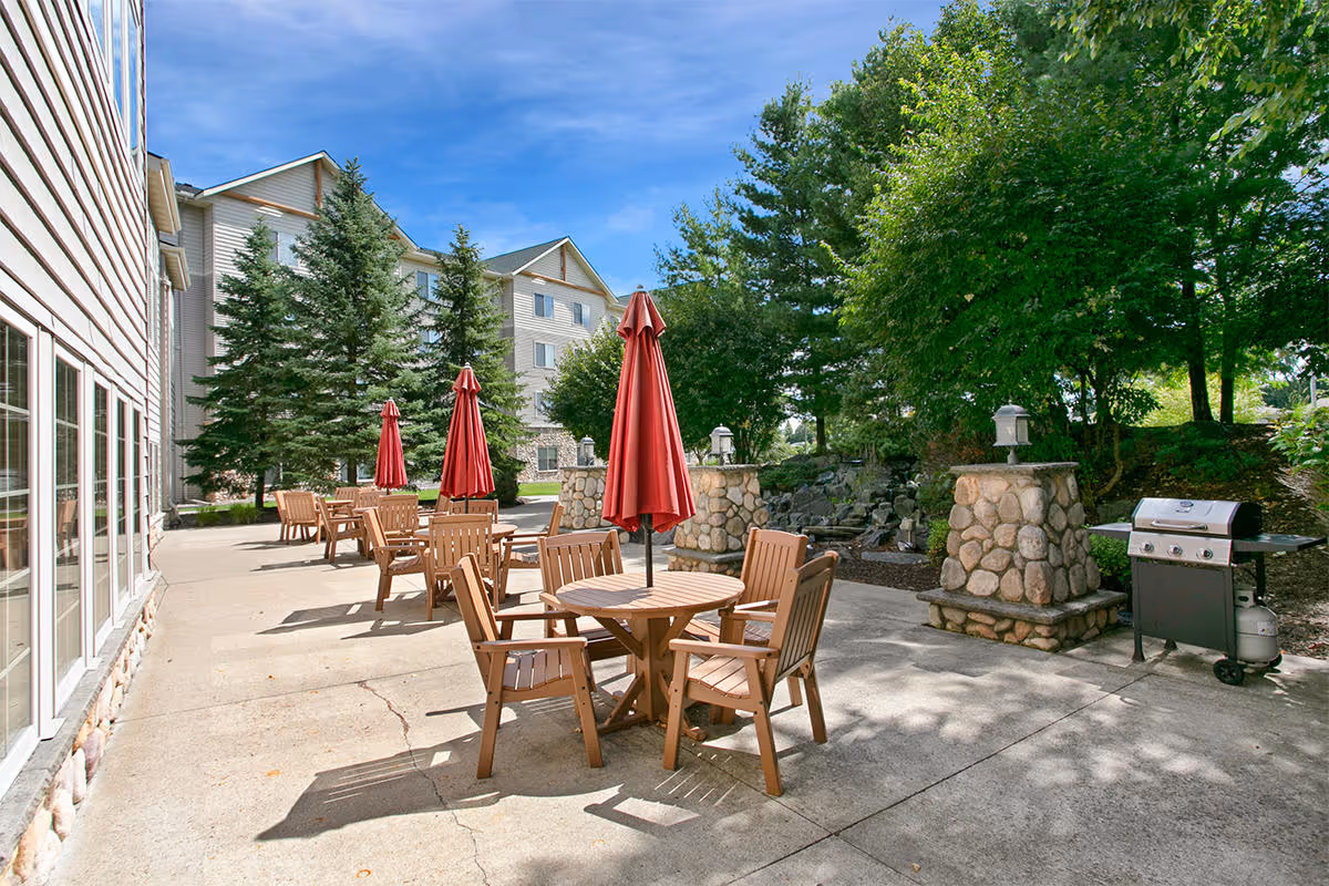 Outdoor patio with wooden tables and red umbrellas, a barbecue grill, and landscaping beside a multi-story building.