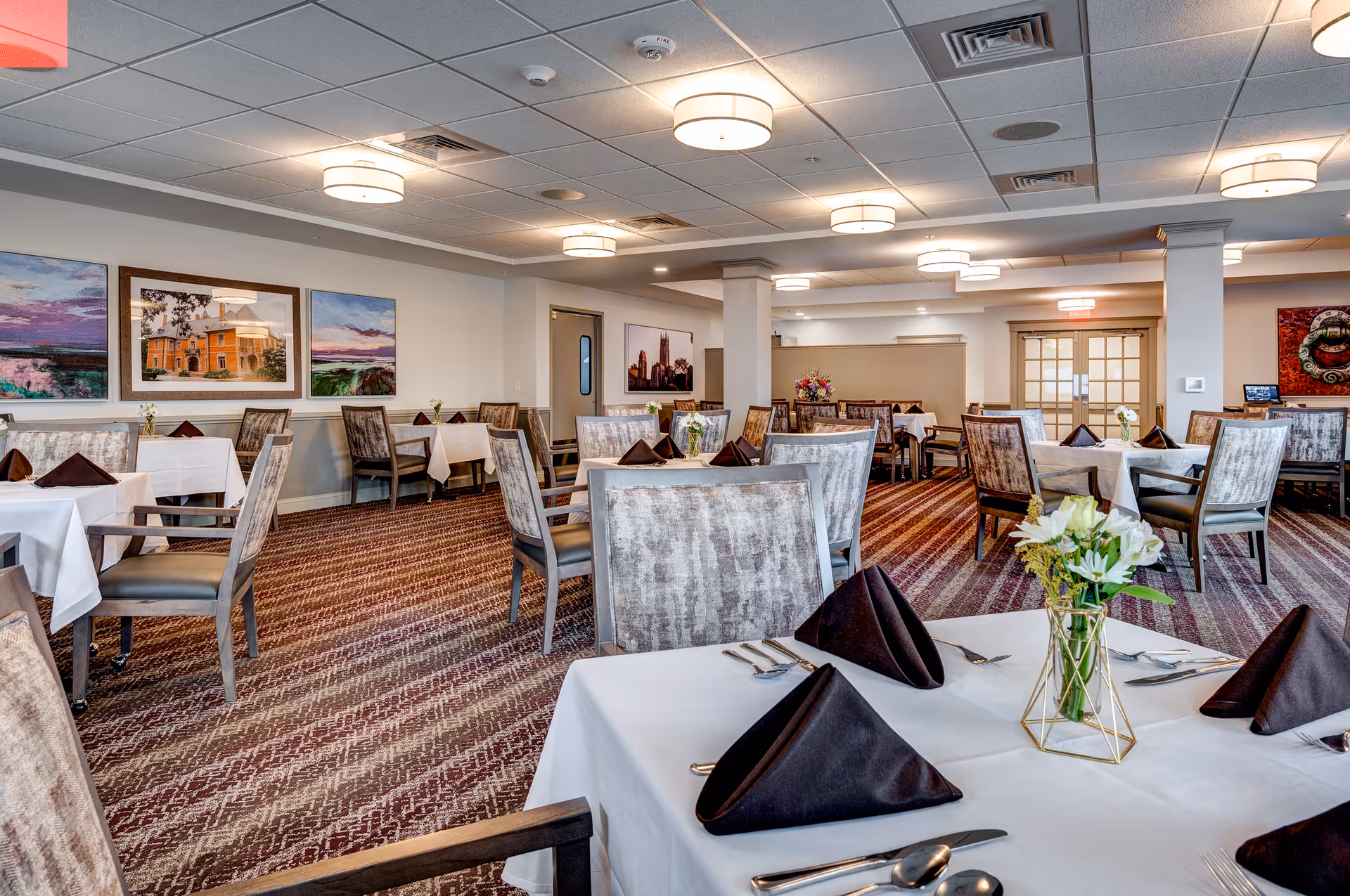 A spacious dining room with multiple tables covered in white tablecloths, each set with dark folded napkins, silverware, and small floral centerpieces. The room features patterned carpet, cushioned chairs, ceiling lights, and framed artwork on the walls.