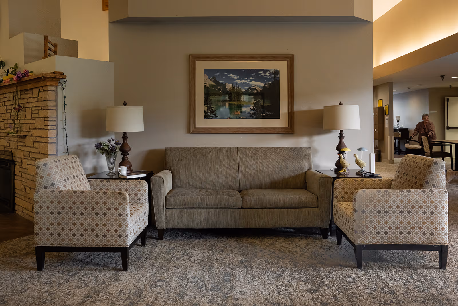 A cozy living room area with a beige sofa flanked by two patterned armchairs. There are two side tables with lamps and decorative items on either side of the sofa. A framed landscape painting hangs on the wall above the sofa. To the left, there is a stone fireplace with some flowers on top. In the background, an elderly person is seen walking in a hallway.