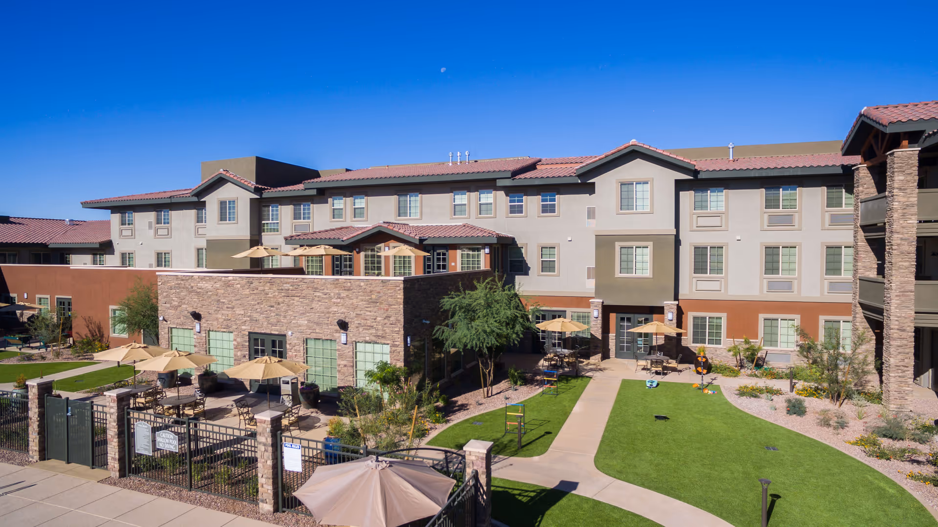 Exterior view of The Enclave at Anthem Senior Living facility showing a multi-story building with beige and brown walls, red-tiled roof, and multiple windows. The foreground features a fenced patio area with tables, chairs, and umbrellas, surrounded by landscaped greenery and a walking path under a clear blue sky.