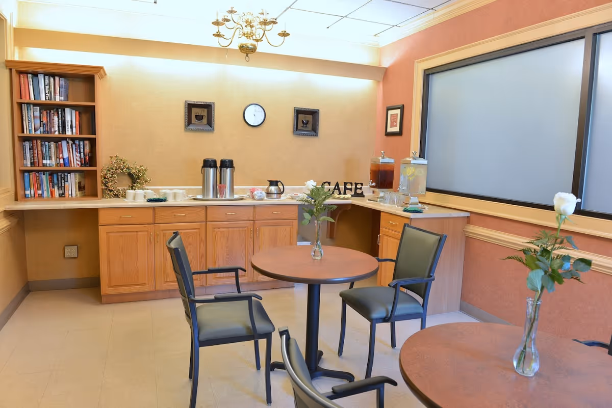 A small cafe area with round tables and chairs, a countertop with coffee dispensers, cups, and beverage containers, a bookshelf filled with books, and decorative flowers in vases on the tables.