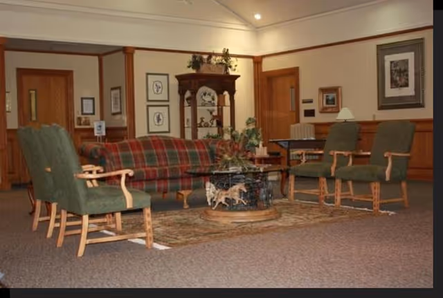 Carpeted lounge area with a plaid sofa, green armchairs arranged around a decorative coffee table and shelving in a senior facility common room.